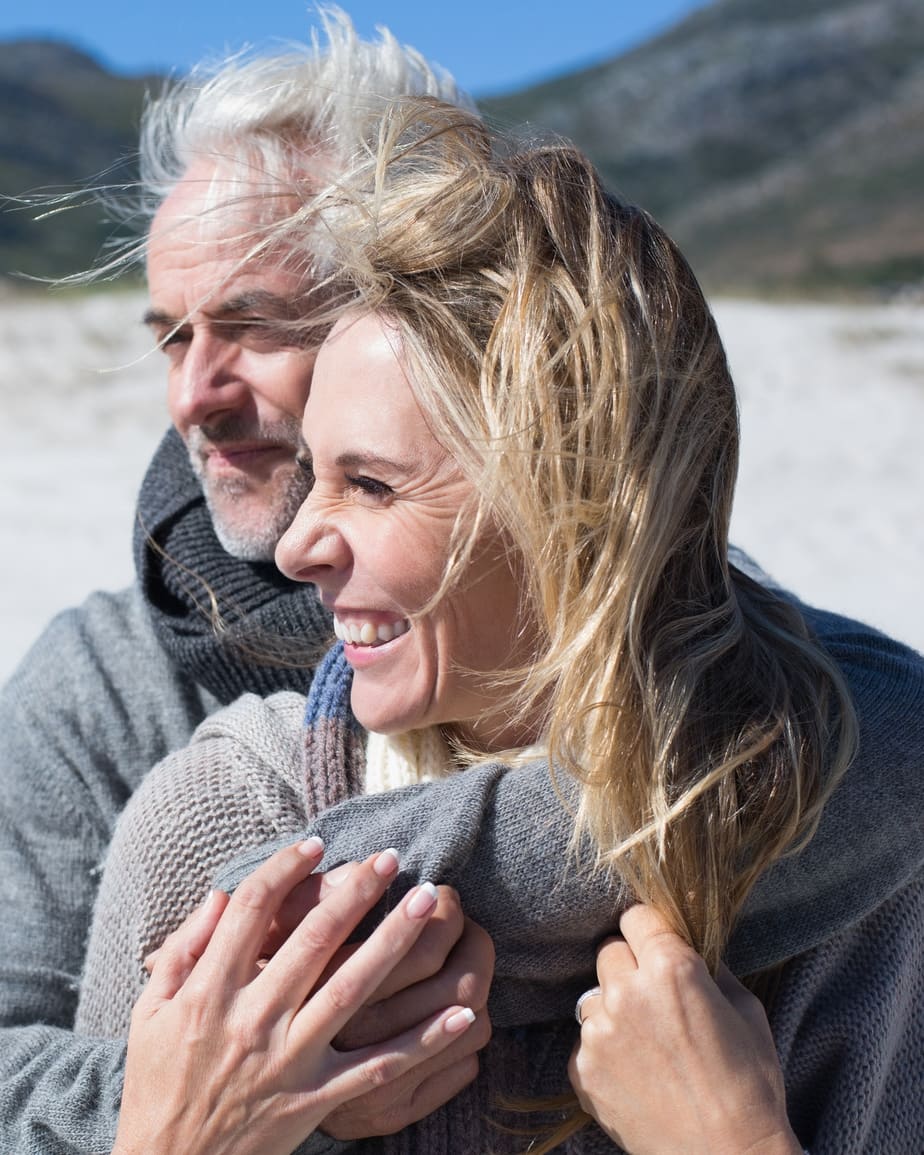 A couple on a beach on a bright cold day, wrapped up warm, his arms around her and both of them laughing