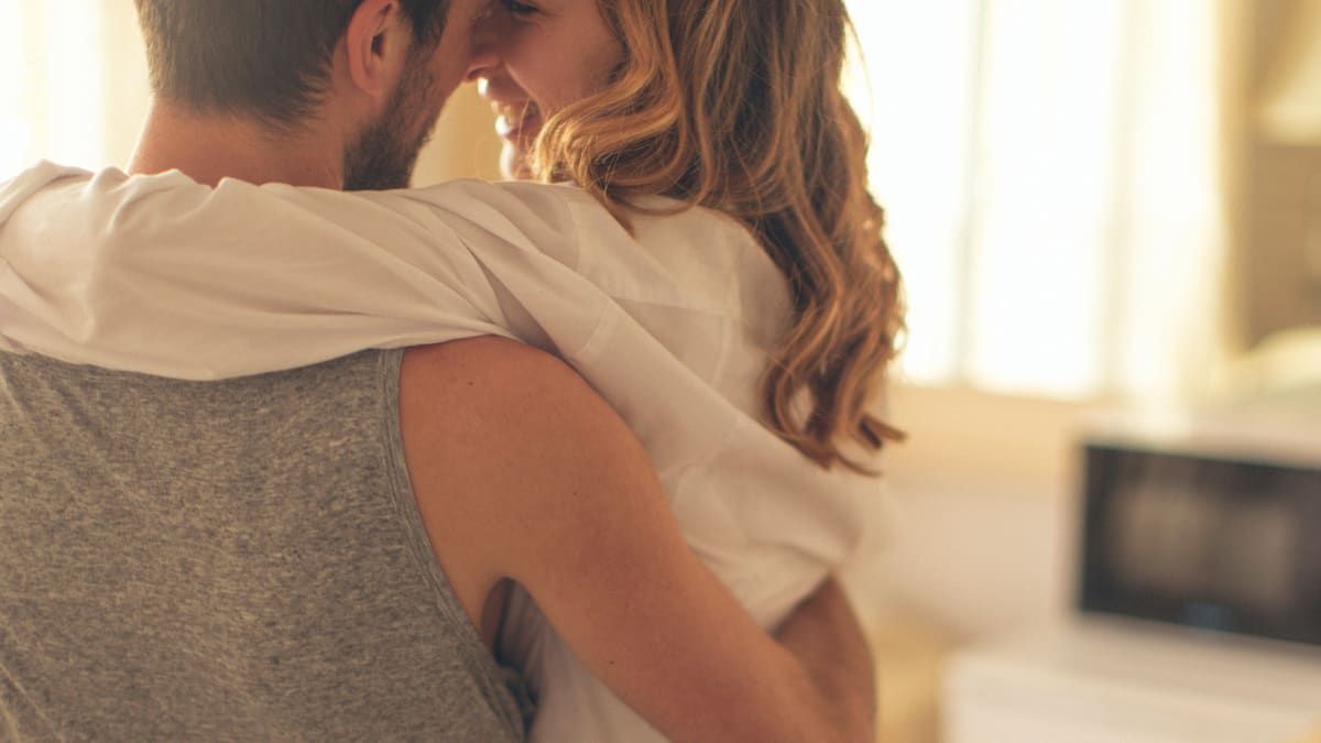 A couple holding each other in a sunlit room at home, both smiling