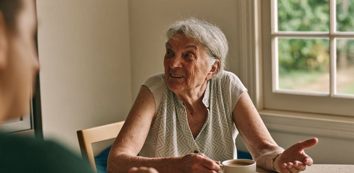 A woman in her seventies at her kitchen table with a cup of tea, mid conversation