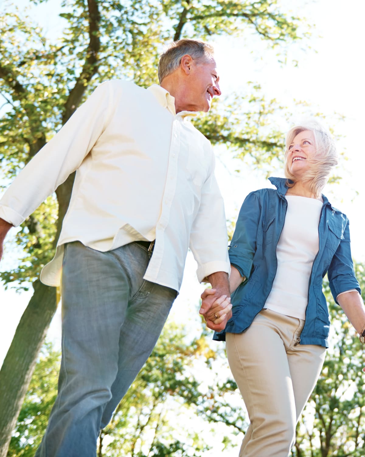 A couple in their sixties walking hand in hand under trees on a bright day