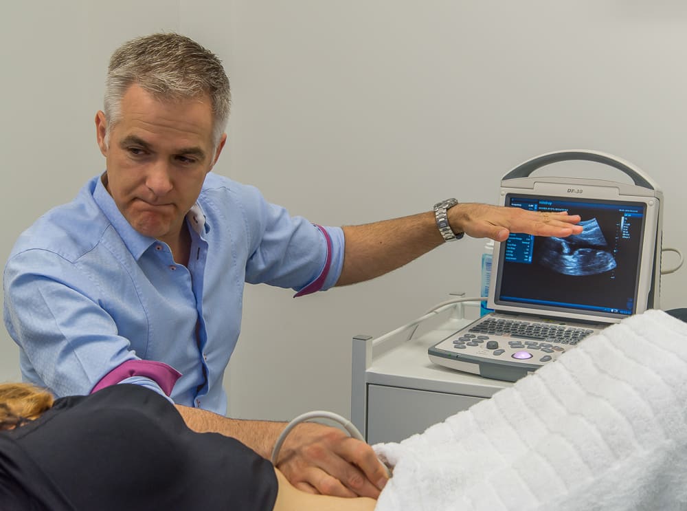 Nathan Scott running a real-time ultrasound scan over a patient's lower abdomen, the image on the screen beside them